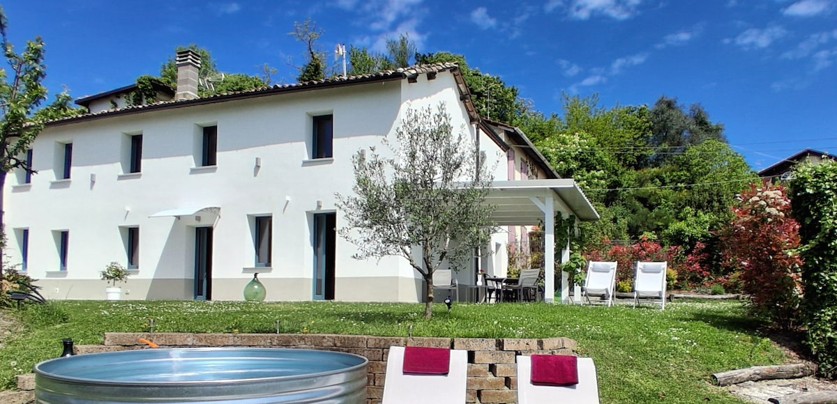 A spacious house is framed by lush greenery, featuring a white exterior that reflects sunlight. A covered patio with dining seating offers comfort, while sun loungers invite relaxation. A circular pool is positioned nearby, surrounded by a well-maintained garden with colorful foliage.