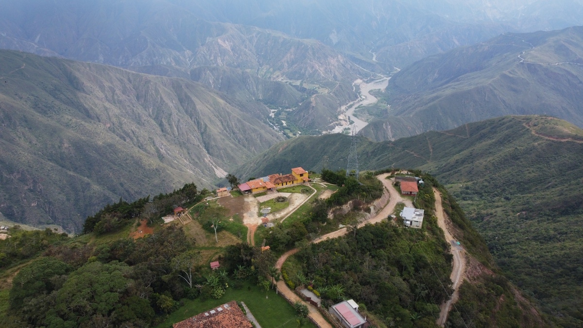 An aerial view captures a serene mountainous landscape featuring hills and valleys with a winding river below. Several chalets are seen nestled among lush greenery, emphasizing the natural surroundings. The layout of paths and fields suggests accessibility and tranquility in this remote location.