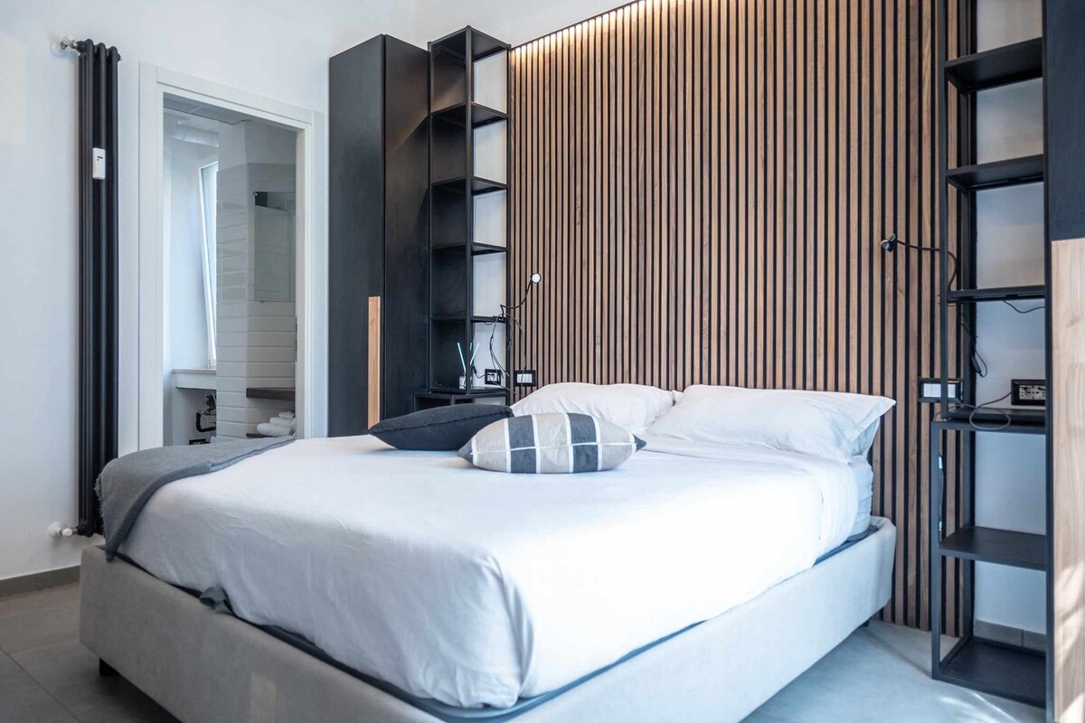 A spacious bed is centered against a wooden slatted feature wall. Flanking the bed are dark shelving units, offering storage and accent lighting, while a soft grey blanket drapes casually. A doorway leads to a well-lit bathroom area in the background.