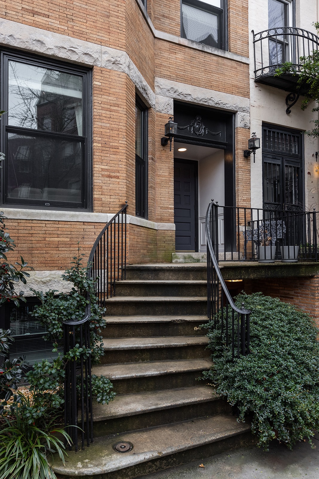 The entrance of the apartment showcases elegant brickwork and a dark door flanked by stylish light fixtures. A set of stone stairs, bordered by lush greenery, leads to the entrance, enhancing the home's inviting exterior.