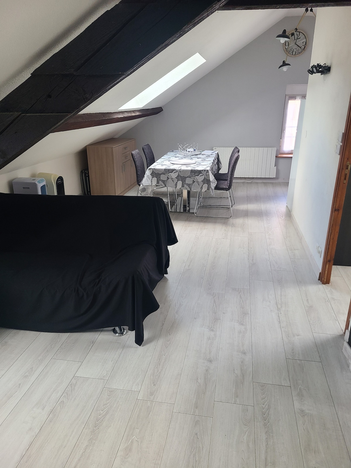 A spacious living area features a dining table set for four and a comfortable sofa. Natural light enters through a skylight, illuminating the light-colored flooring. A storage cabinet is positioned along one wall, contributing to the open ambiance of the space.