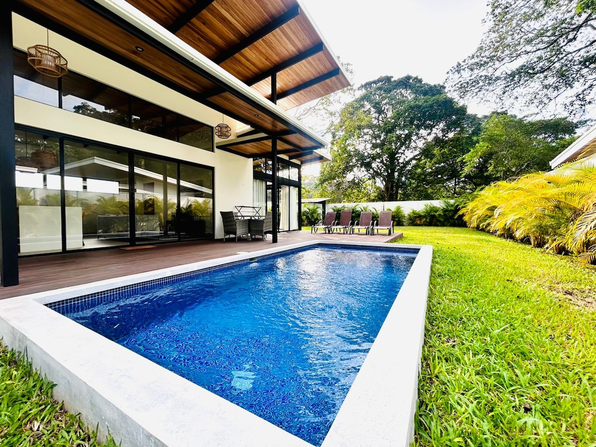 An inviting outdoor area features a sparkling blue pool bordered by lush green grass. A spacious wooden deck provides access to the house, with sleek glass doors and comfortable seating options nearby. Tall trees surround the space, creating a peaceful atmosphere.