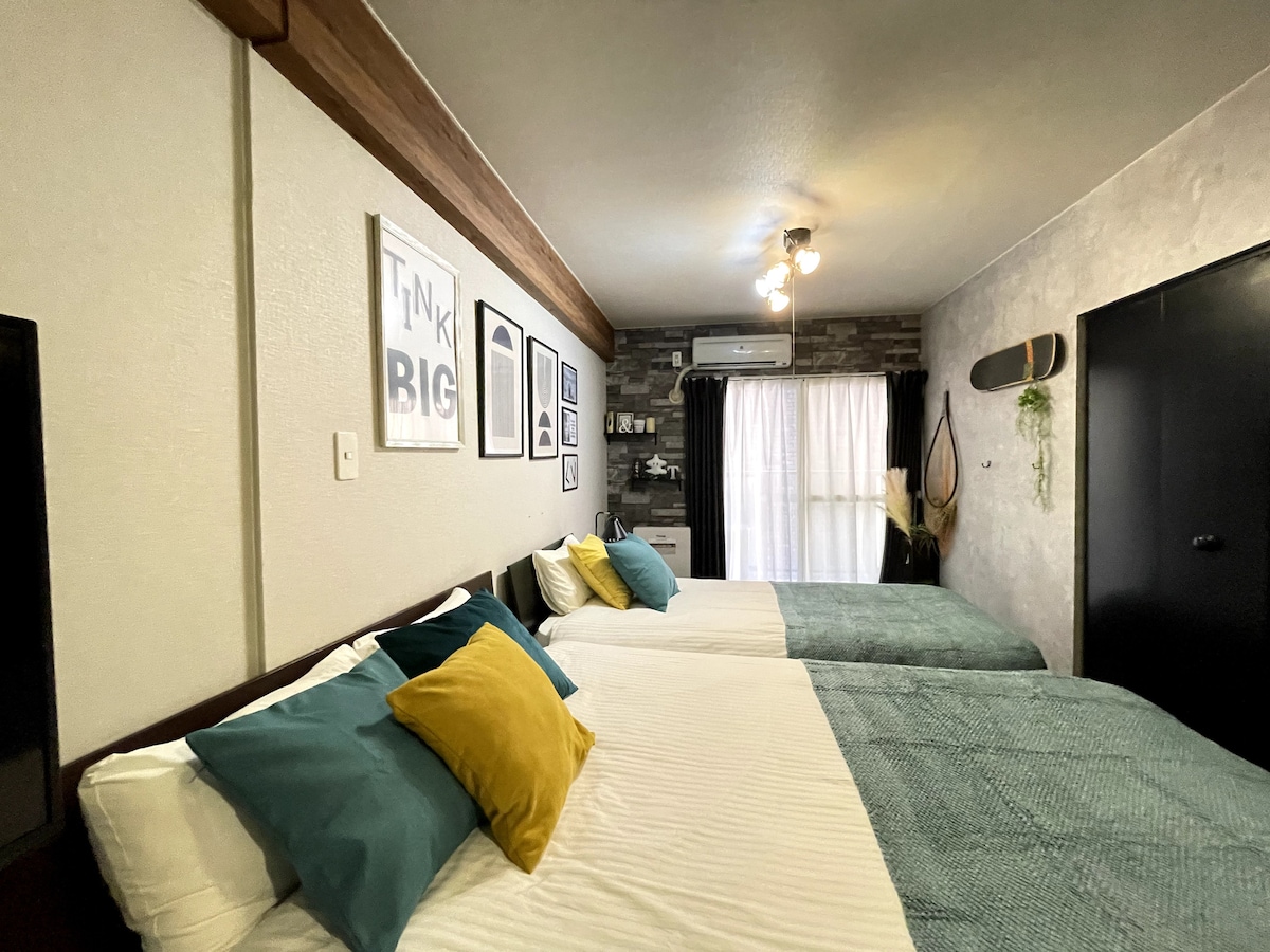 The bedroom features two spacious beds dressed in soft linens. Colorful pillows in teal and yellow add a lively touch. A window covered by sheer curtains allows natural light to brighten the room, while wall art and decorative elements provide visual interest.