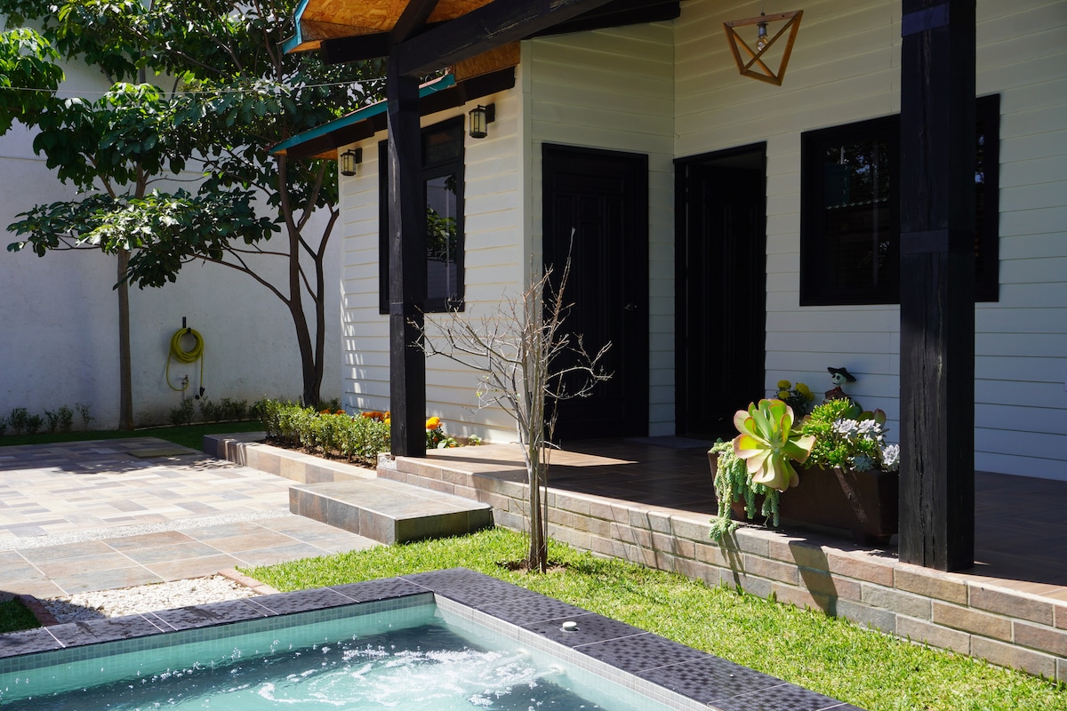A charming exterior view is presented, featuring a well-kept porch with black accents and a small landscaped garden. A cozy hot tub, surrounded by smooth tiles, is visible in the foreground, reflecting the greenery of nearby plants and trees.