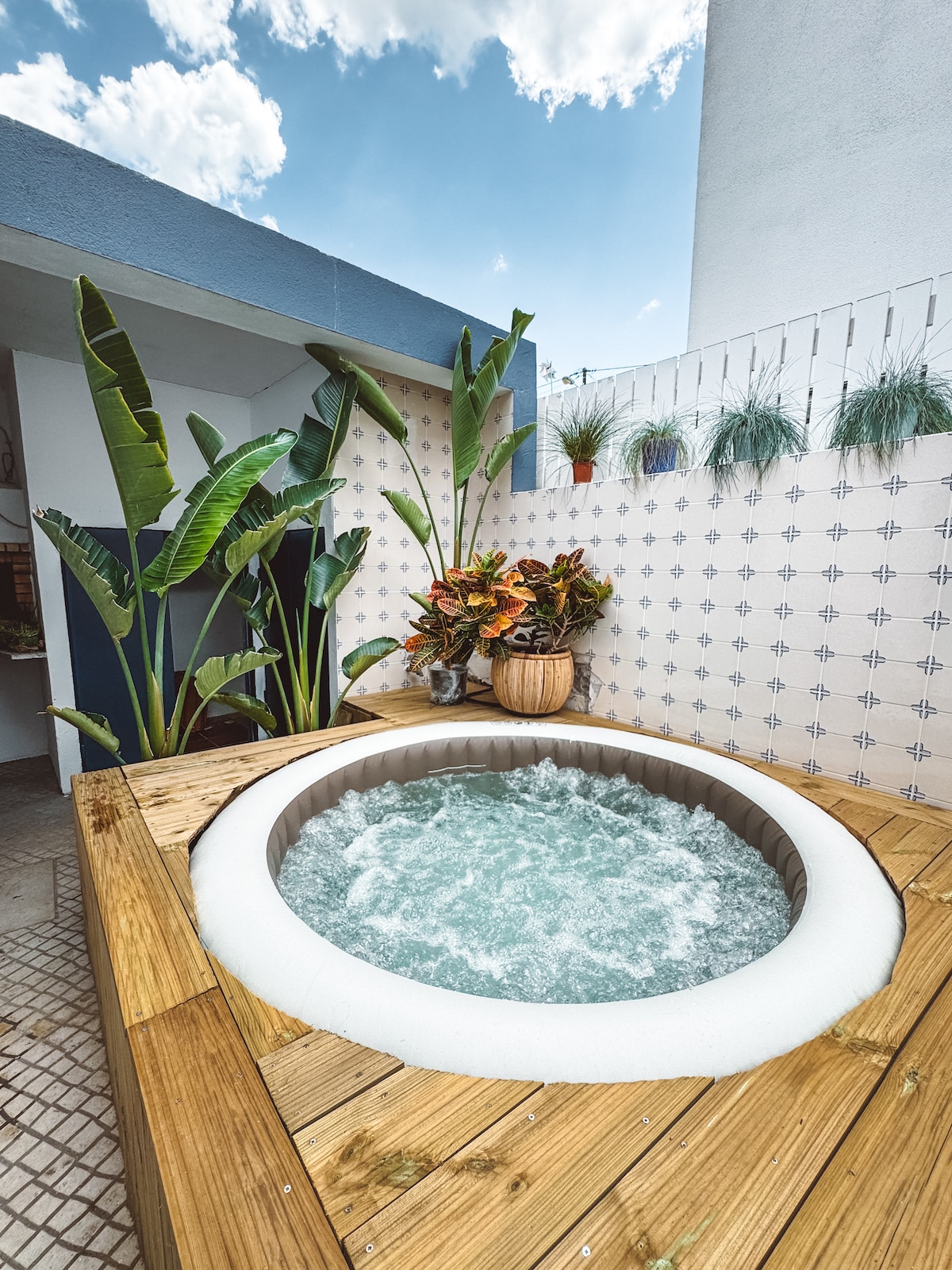 A hot tub is surrounded by wooden decking, creating a relaxing outdoor space. Lush green plants, including tall banana leaves and colorful potted flowers, are displayed nearby. A textured tiled wall adds visual interest while the sky is partly cloudy, allowing natural light to brighten the area.