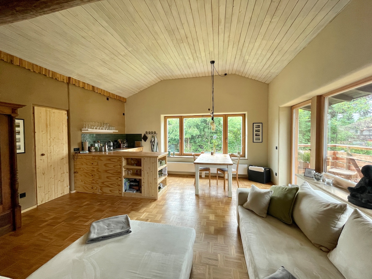 A spacious living area is showcased with a wooden ceiling and large windows that offer views of greenery. A dining table is set in the center, while an open kitchen features natural wood cabinetry. A comfortable sofa is positioned nearby, inviting relaxation.