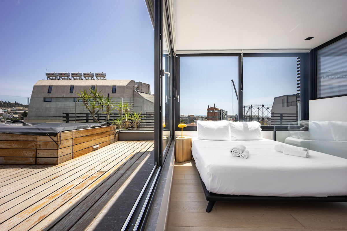 A bright bedroom features floor-to-ceiling windows, offering clear views of the cityscape. The spacious bed is dressed in crisp white linens. A wooden deck extends outside, complemented by greenery and modern outdoor furnishings. Sunlight floods the room, creating an open and airy ambiance.