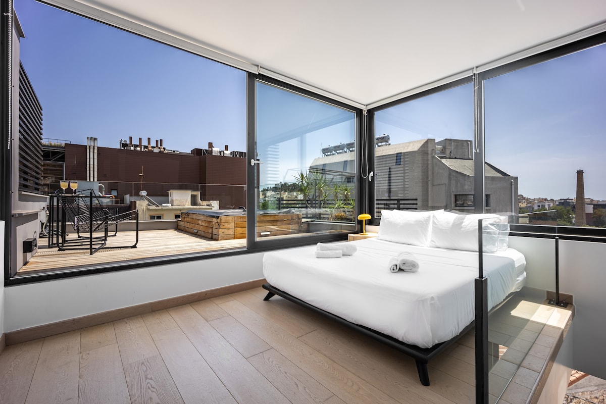 A spacious sleeping area is highlighted by large floor-to-ceiling windows allowing ample natural light. A comfortable bed is made with crisp white linens. Outside, a deck is visible, furnished with seating and offering city views.