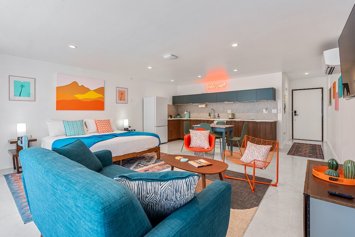 A bright open living space showcases a spacious bed against a backdrop of desert-themed artwork. Comfortable seating includes a blue sofa and orange chairs. A dining area is seen, accompanied by a kitchen with modern appliances, while soft lighting enhances the welcoming atmosphere.