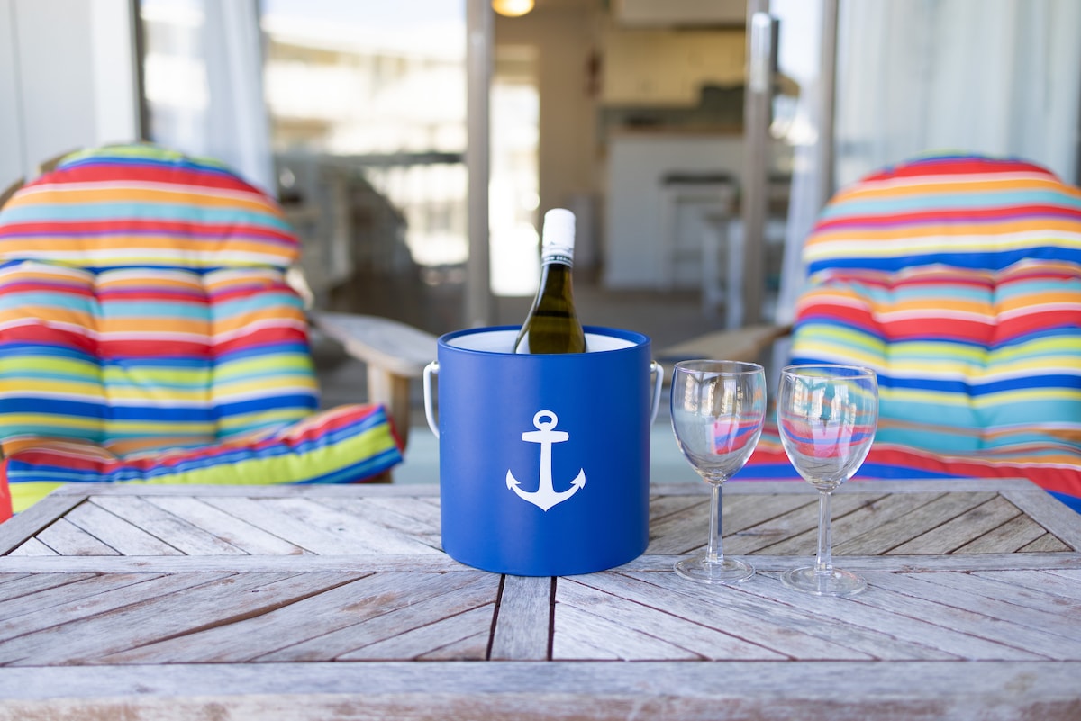 A vibrant outdoor seating area features two colorful, striped lounge chairs positioned beside a wooden table. An ice bucket with a wine bottle and two glasses is centered on the table, inviting relaxation and enjoyment. The backdrop reveals an open living space.