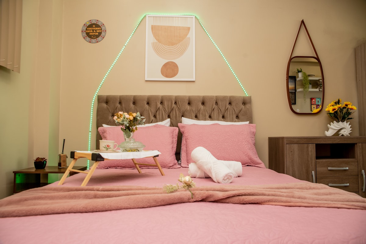 A spacious bed is dressed in pink linens, featuring decorative pillows and a soft throw. Small trays with tea and flowers are placed on top. A mirror and wall art are positioned above the bed, while ambient lighting adds a gentle glow to the area.
