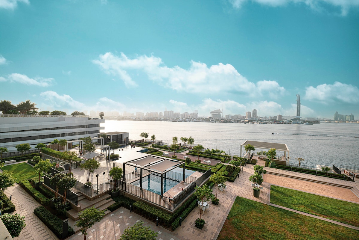 An expansive view of a well-maintained outdoor space featuring neatly arranged greenery and walking paths. A swimming pool is visible in the foreground, with the vibrant backdrop of the city skyline and waterway under a clear blue sky.