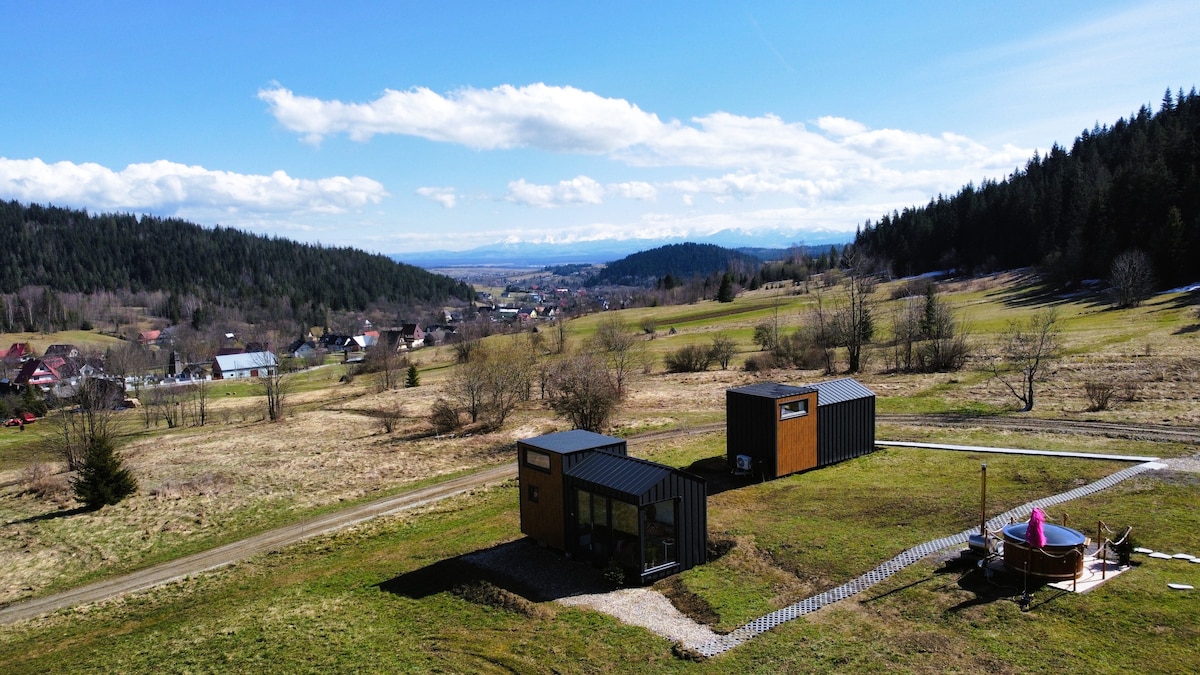 A spacious landscape is featured, showcasing two distinct tiny houses placed among lush greenery and rolling hills. The background reveals distant mountains under a bright blue sky with scattered clouds. A pathway leads toward the houses, enhancing the serene natural setting.