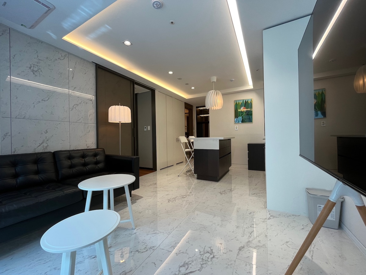 A modern living area features a black leather sofa and two white coffee tables on polished marble flooring. Soft lighting is provided by a pendant lamp overhead, while a flat-screen TV is positioned on an easel. A kitchen area with bar seating is visible in the background.