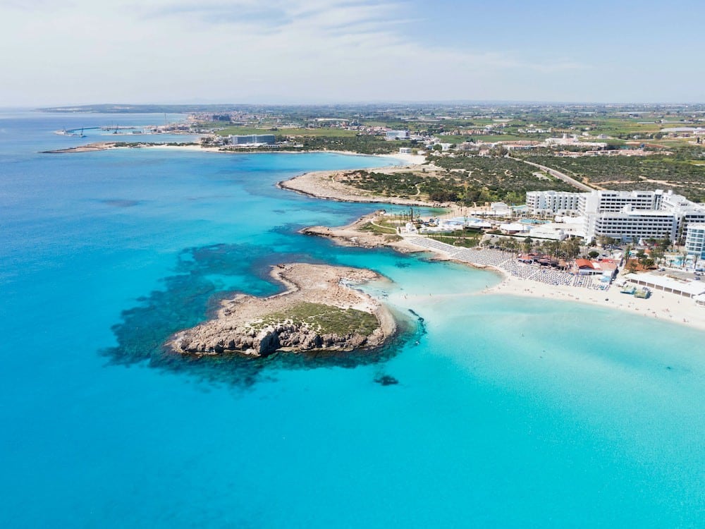 Aerial view showcases the vibrant turquoise waters alongside a sandy beach. The coastline features rocky outcrops and well-developed beachfront properties, with greenery visible in the background. The horizon is highlighted by distant landforms under a clear sky.