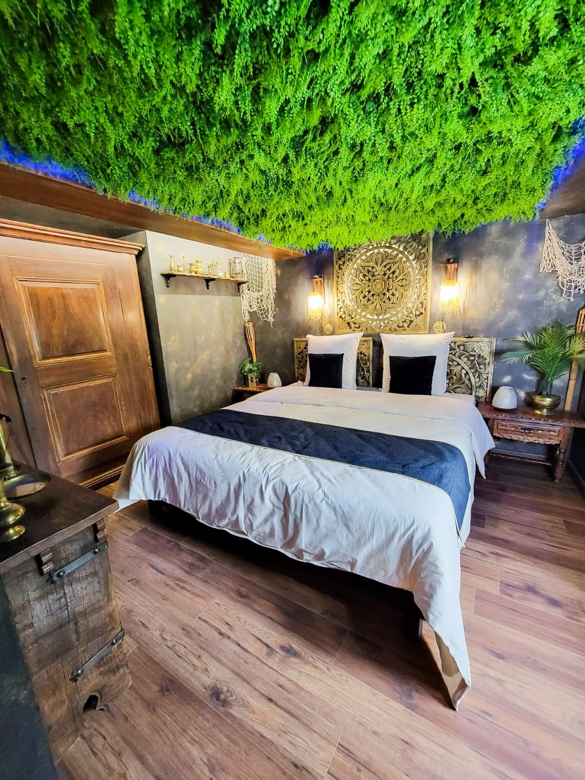 A unique bedroom features a ceiling adorned with lush greenery, enhancing the room's immersive theme. The spacious bed is dressed in crisp white linens with dark accent pillows. Decorative accents include intricate wall art and soft lighting, creating a tranquil environment.