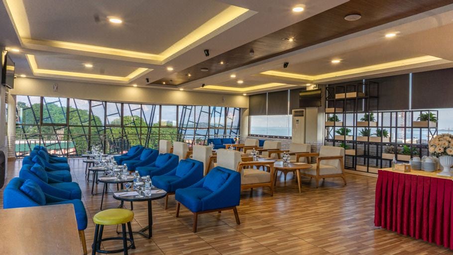 A spacious lounge area features large windows offering scenic views. Colorful blue armchairs surround glass-topped tables, creating a welcoming space for relaxation. A reception area is situated at the back, complemented by decorative plants on shelves, contributing to the overall inviting ambiance.