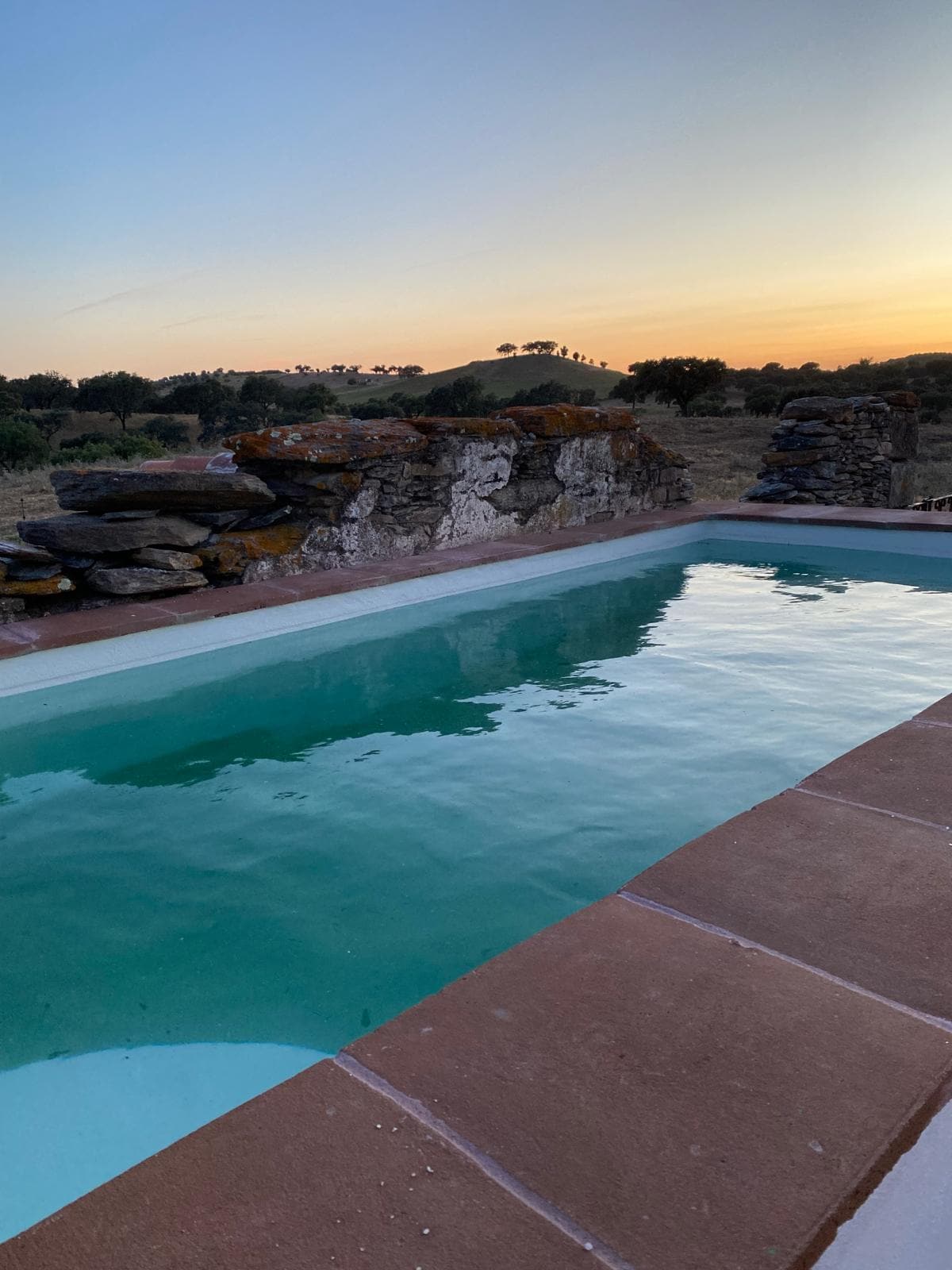 A swimming pool is set against a backdrop of rocky walls, reflecting the colors of the sunset. Surrounding landscapes feature rolling hills and natural vegetation, creating a serene atmosphere for relaxation.