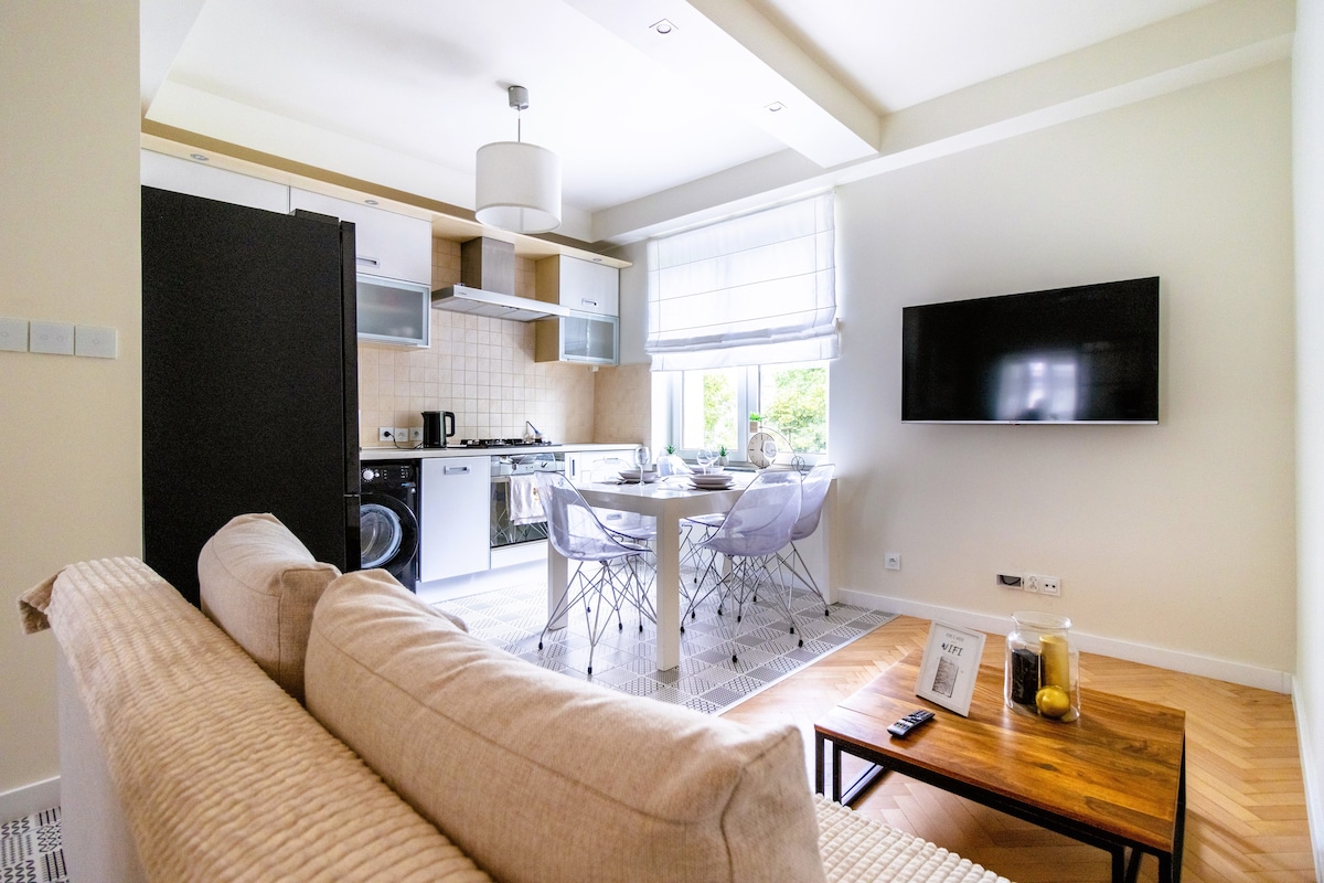 A bright and open kitchenette is visible, equipped with modern appliances including a refrigerator and oven. A cozy living area features a light-colored sofa facing a wall-mounted television. A dining table with chairs is set by the window, allowing for natural light to fill the space.