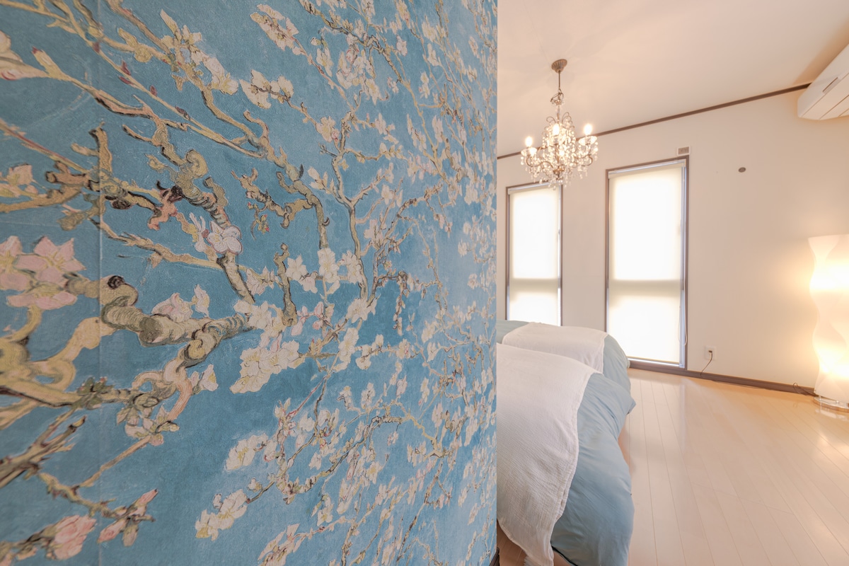 A tranquil bedroom features ornate wallpaper with floral designs in soft hues. A pair of spacious beds dressed in light bedding is positioned across from large windows, allowing natural light to filter in. A contemporary chandelier adds subtle brightness to the room.