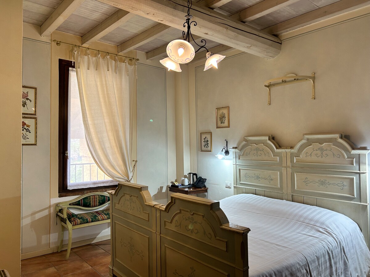 A cozy bedroom displays a light-colored wooden bed frame with decorative detailing. Natural light enters through a window, adorned with a white curtain. A small seating area and a wall-mounted light provide functional space, while decorative artwork adds subtle charm.