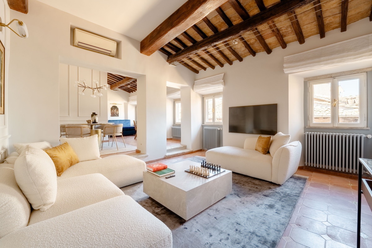 A spacious living area features soft, plush seating arrangements in neutral tones, complemented by a modern coffee table. Large windows allow natural light to fill the space, highlighting the wooden beam ceiling. A sleek television is mounted on the wall, enhancing the relaxed ambiance.