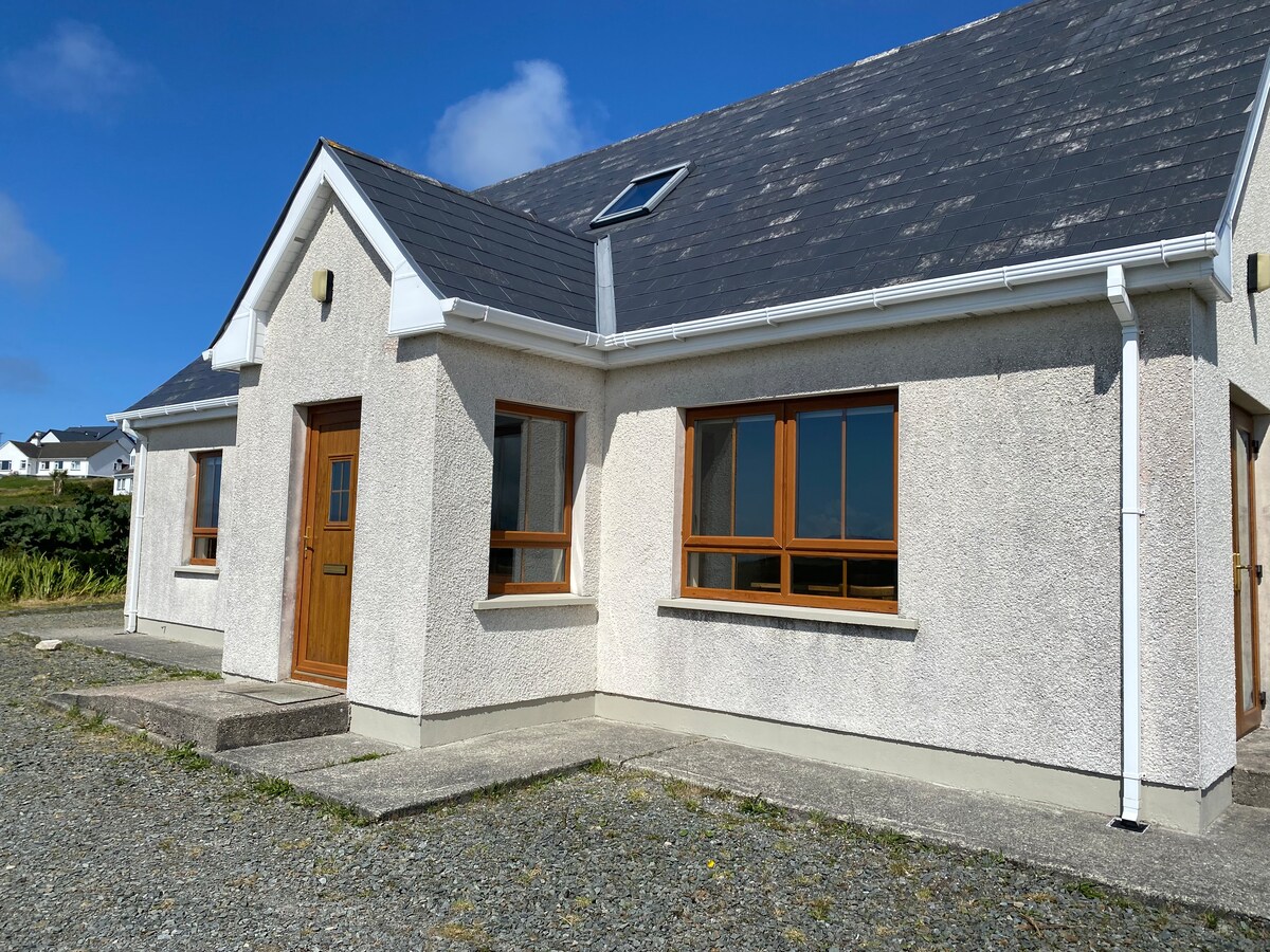A light-colored exterior of a holiday home is presented, featuring multiple windows that allow natural light. The entrance is marked by a wooden door, and stone steps lead to the front porch. A clear blue sky is visible above, enhancing the serene outdoor setting.