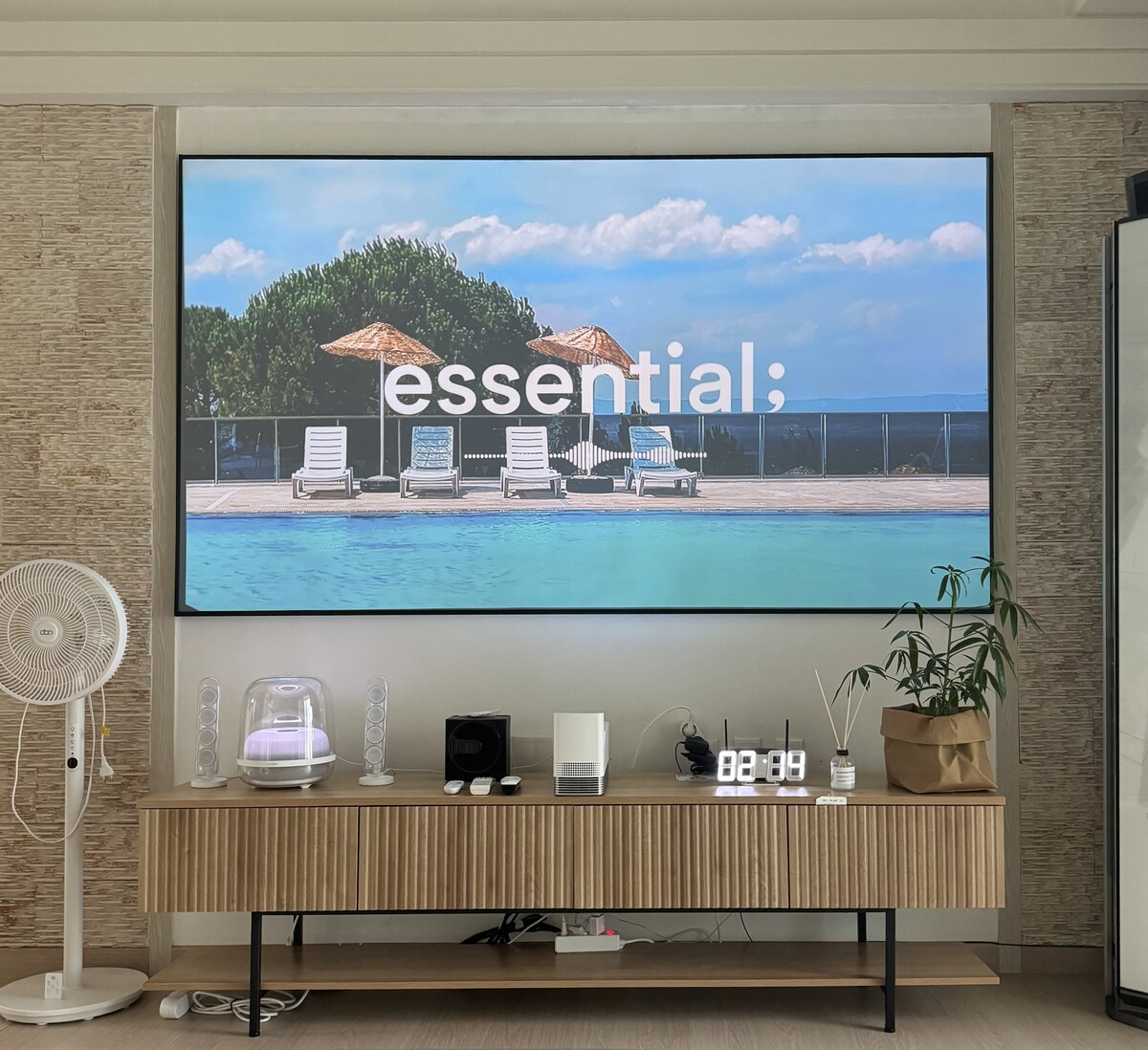 A large screen displays a serene pool area with lounge chairs and two parasols under a clear blue sky. A sleek wooden console holds decorative items, a fan, and a digital clock. The atmosphere is calm and inviting.
