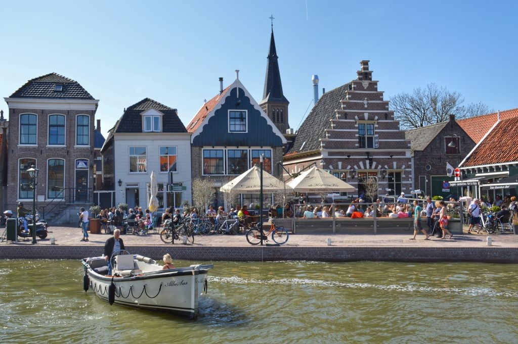 A scenic waterfront view features historic buildings with distinct architecture. Outdoor seating areas are occupied by guests enjoying the atmosphere. A small boat glides along the calm water, complementing the lively scene of bicycles parked nearby and people socializing along the promenade.