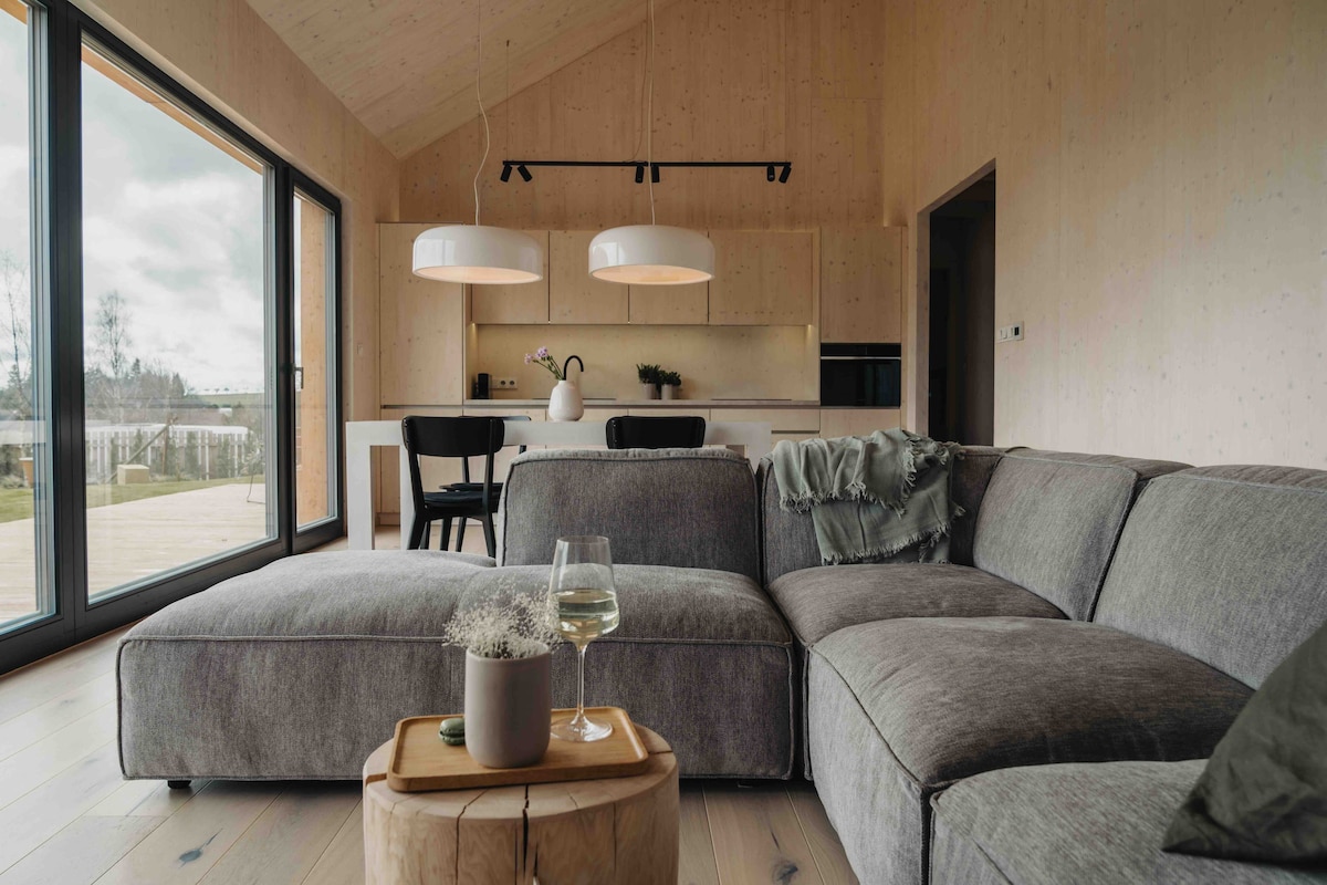 A spacious living area features a comfortable sectional sofa in muted tones. Large glass doors provide a view of the outdoor space. A wooden side table holds a glass of wine and a small vase with herbs. The modern kitchen and dining area can be seen in the background.