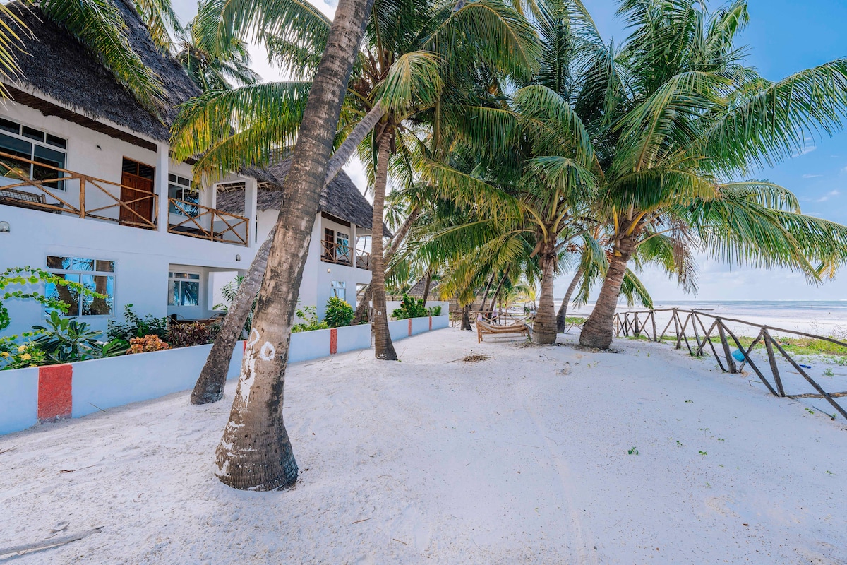 The exterior of the duplex bungalow is surrounded by lush palm trees, with a pathway of soft white sand leading towards the beach. The building features a traditional thatched roof, and spacious outdoor areas are visible, ideal for relaxing and enjoying the serene coastal environment.