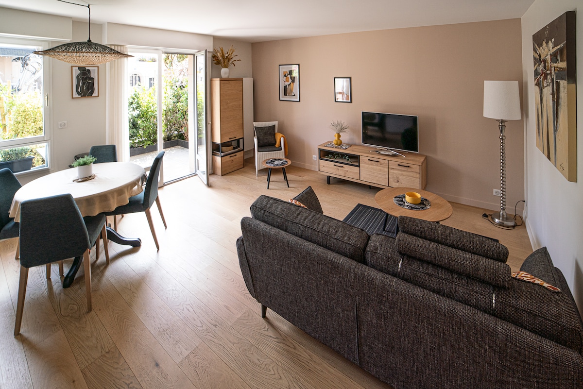 A bright living area features large windows that open onto a green outdoor space. A comfortable couch with an accent pillow faces a media console with a television. A round dining table for four sits beside the couch, accompanied by modern chairs, creating an inviting space.