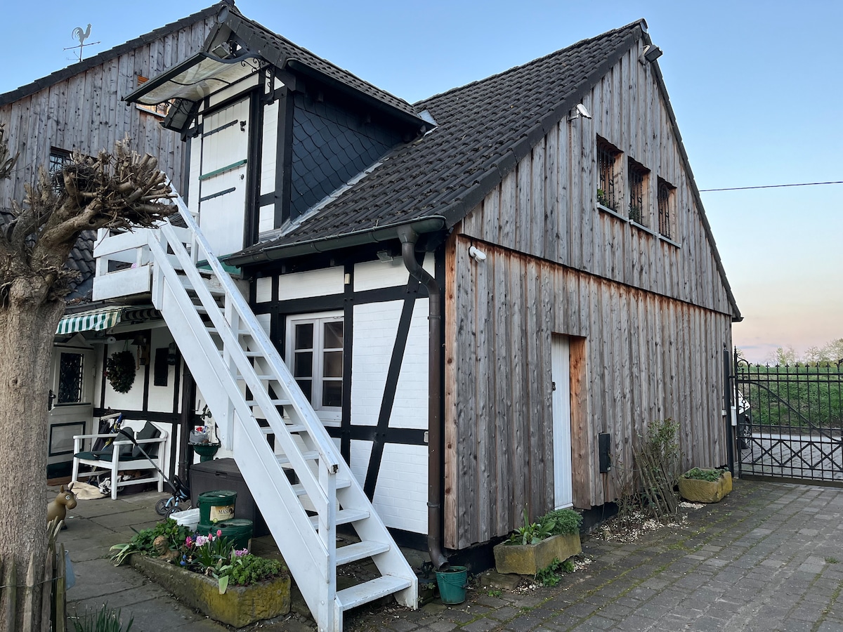 A rustic wooden exterior of a charming building is showcased, featuring a separate entrance with white stairs leading to the upper level. The structure is adorned with greenery and subtle landscaping elements, while the traditional frame design complements its unique character.