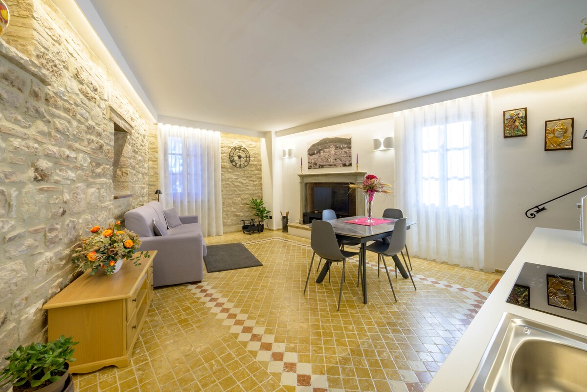A cozy living area features stone walls and soft lighting, creating a warm ambiance. A light purple sofa is positioned next to a dining table with four chairs. A fireplace is visible along with decorative plants near windows dressed in sheer curtains, allowing natural light to fill the space.