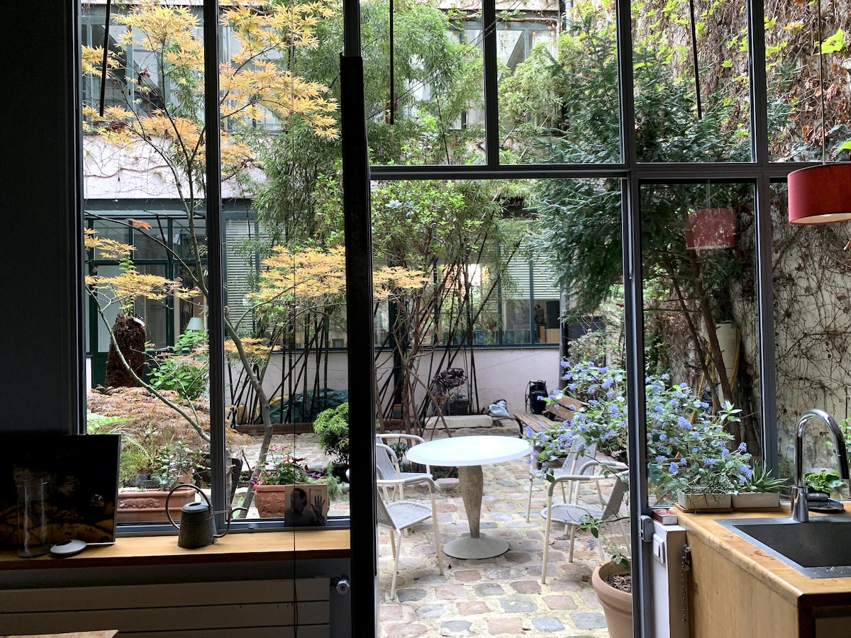 A serene courtyard is viewed through large glass windows, featuring a round dining table surrounded by several chairs. Lush greenery, including bamboo and flowering plants, creates a peaceful atmosphere, while natural stone pathways add texture to the outdoor space.