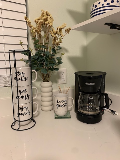 A coffee-making corner is displayed, featuring a coffee maker beside a decorative mug with the words 'be our guest.' Three stacked mugs with motivational phrases are positioned next to a white vase containing dried flowers. Soft light enters through a nearby window.