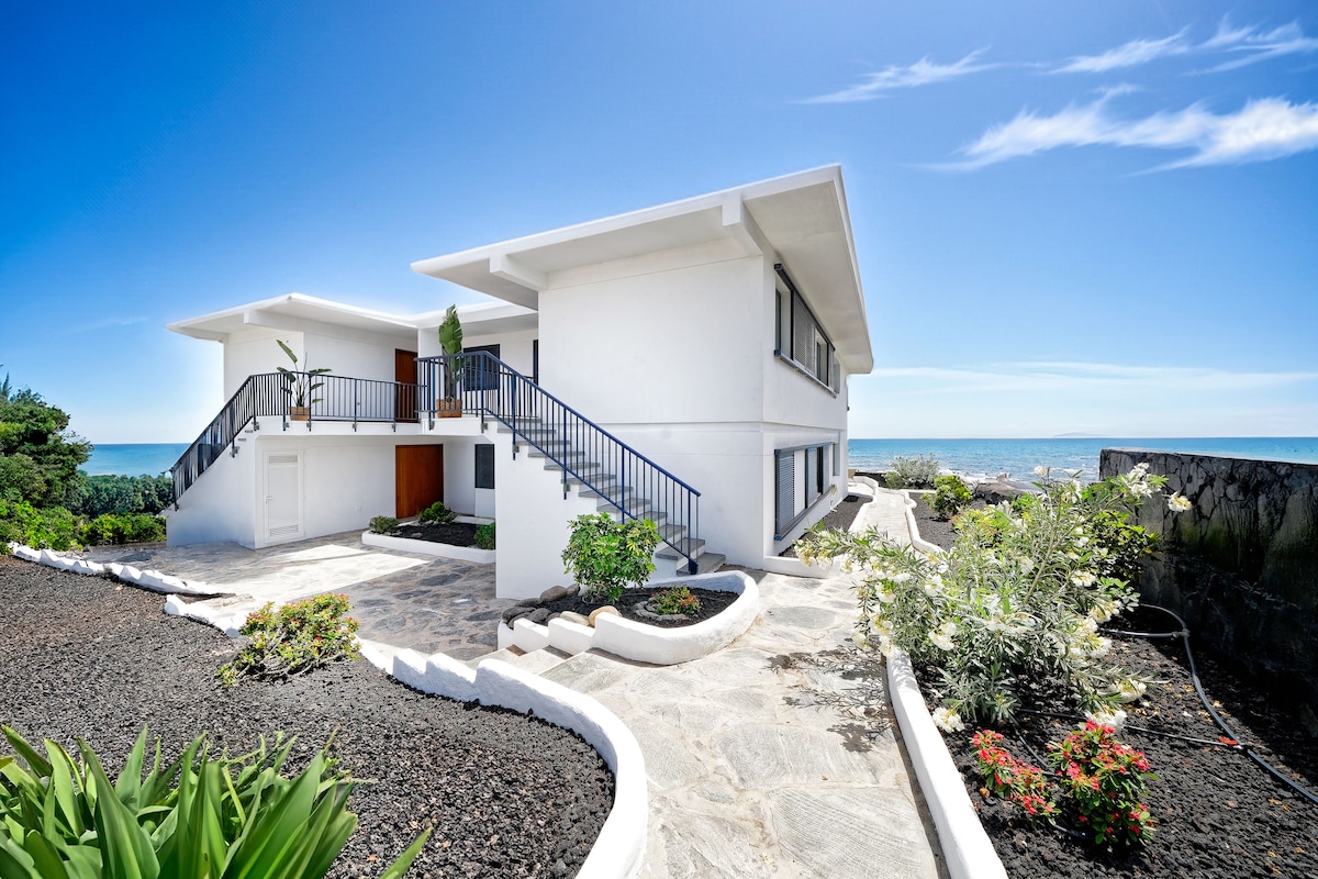 The exterior of the apartment is showcased, featuring a two-story white building with a flat roof. A stone pathway winds through landscaped gardens with native plants and flowers, leading to the entrance. The ocean can be seen in the background under a clear blue sky.
