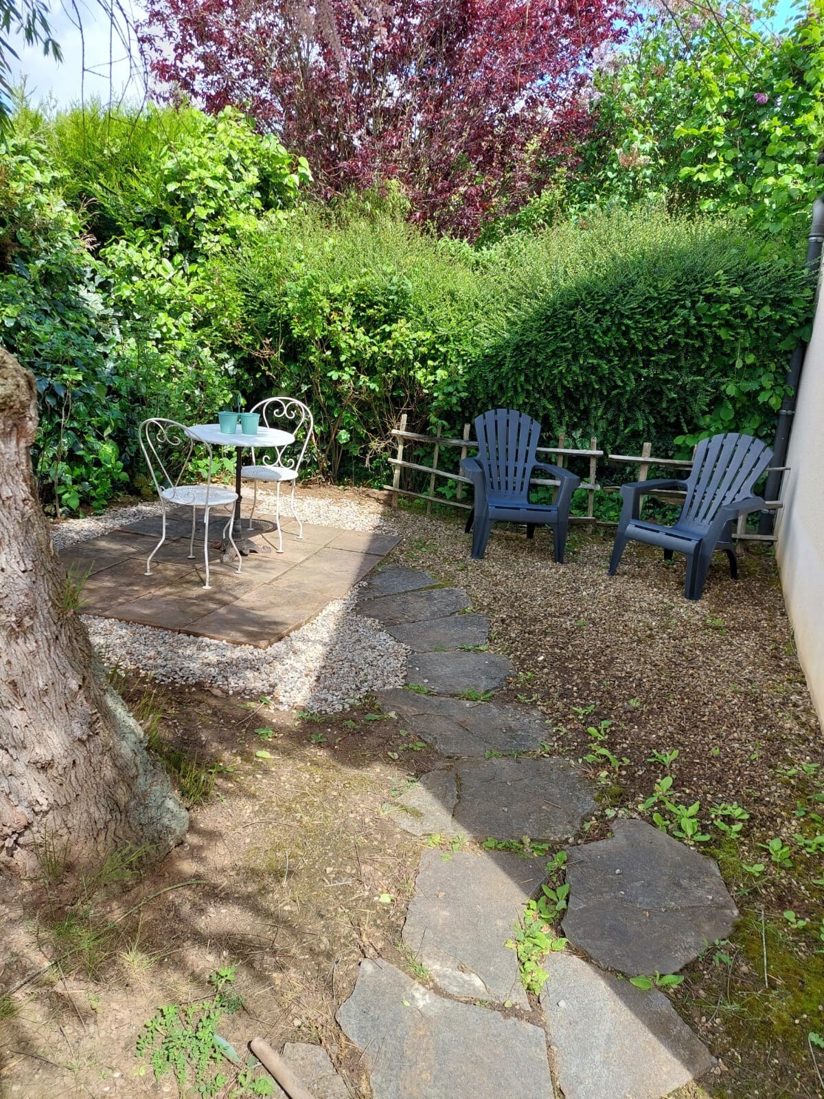 A small outdoor space features a stone pathway leading to a seating area. A round table with two chairs is positioned on a gravel surface, surrounded by lush greenery. Two Adirondack chairs offer additional seating against the backdrop of vibrant foliage.