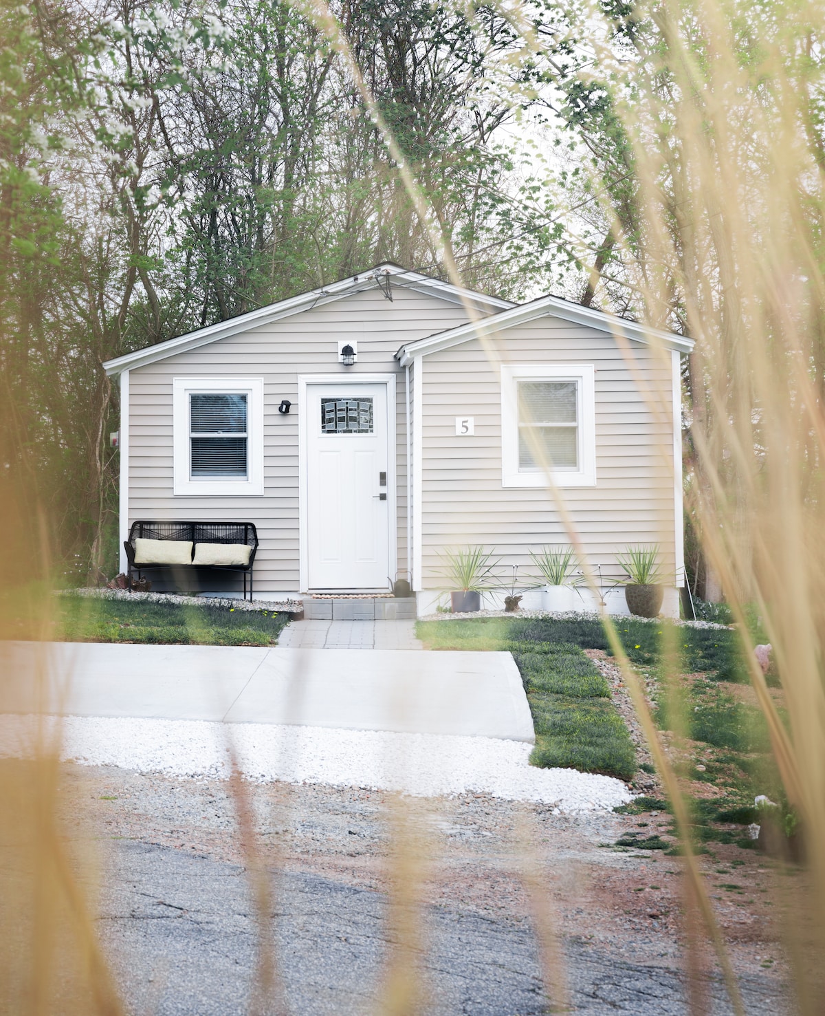 The exterior of the bungalow is visible with a light-colored facade and a welcoming front door. A small bench sits on the front porch, and neatly trimmed plants frame the entrance. A driveway leads to the structure, surrounded by a green lawn and trees.