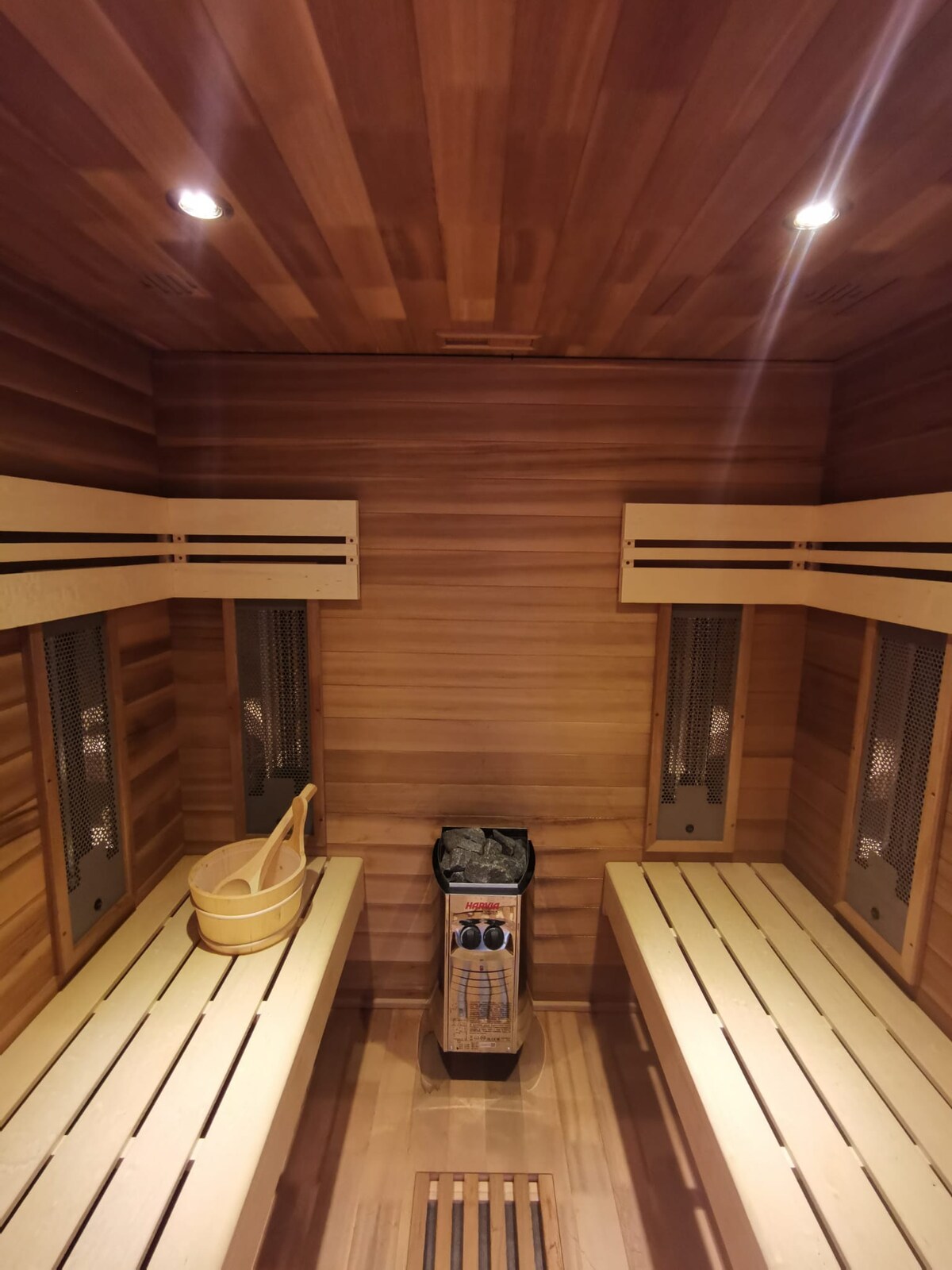 A warm wooden sauna is presented with two long, simple benches on each side, allowing for comfortable seating. A wooden bucket rests on one bench, and a heater is centered at the back, contributing to a serene and calming environment.