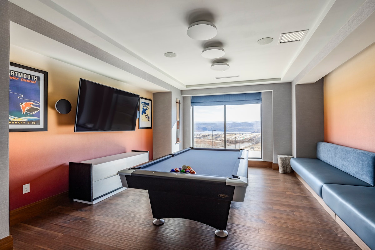 A game room features a billiards table with a full set of colorful balls. A large television is mounted on the wall, and a comfortable blue seating area provides a relaxing space. Scenic views are visible through the expansive window, complemented by softly lit ceiling fixtures.