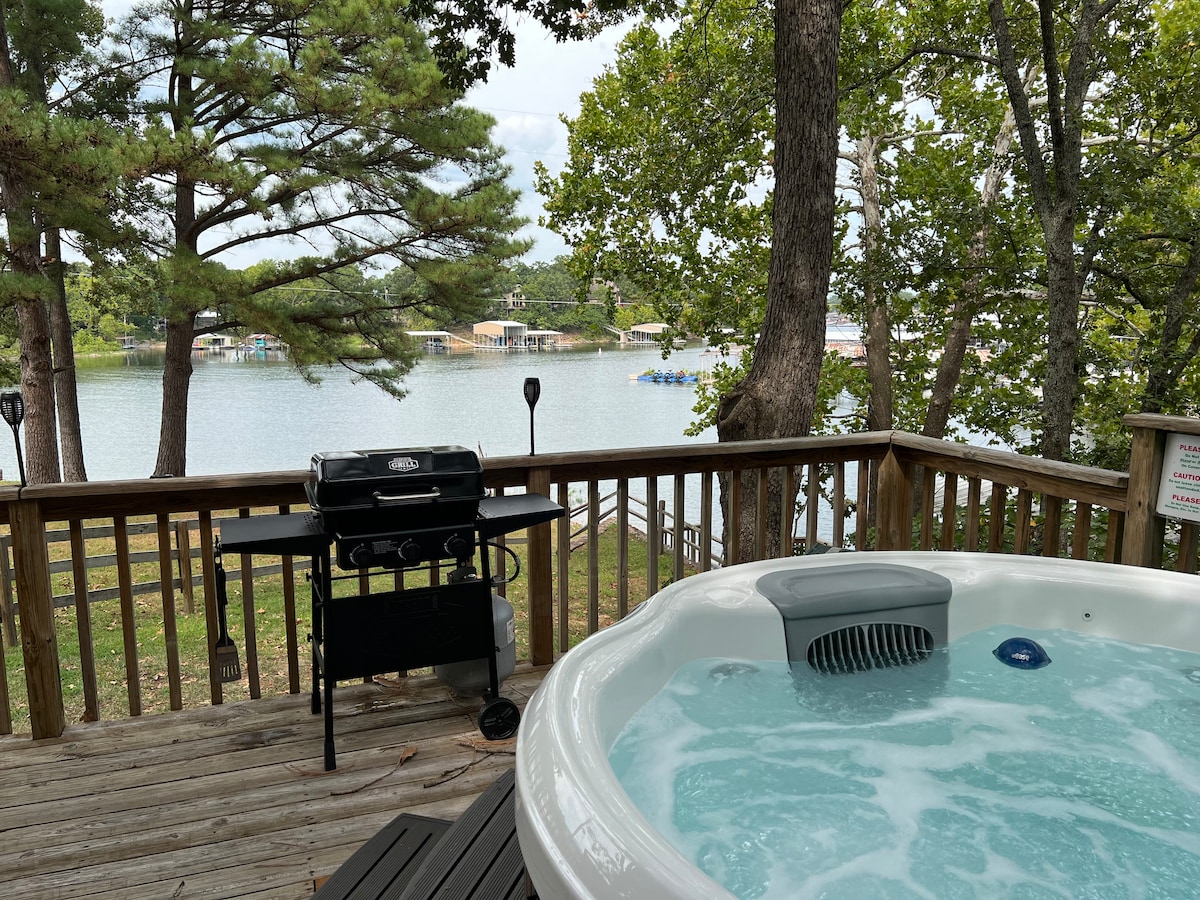 A hot tub sits on a wooden deck, surrounded by trees and overlooking a serene lake. A gas grill is positioned nearby, with seating areas visible on the deck. The sparkling water reflects the greenery, creating a peaceful outdoor setting.