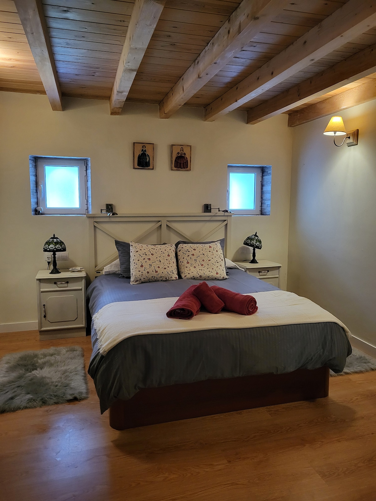 A cozy bedroom features a spacious bed with decorative pillows and rolled towels neatly placed on top. Soft rugs lie on the wooden floor, while warm light is provided by a wall-mounted lamp. Two small windows allow natural light to filter into the room.