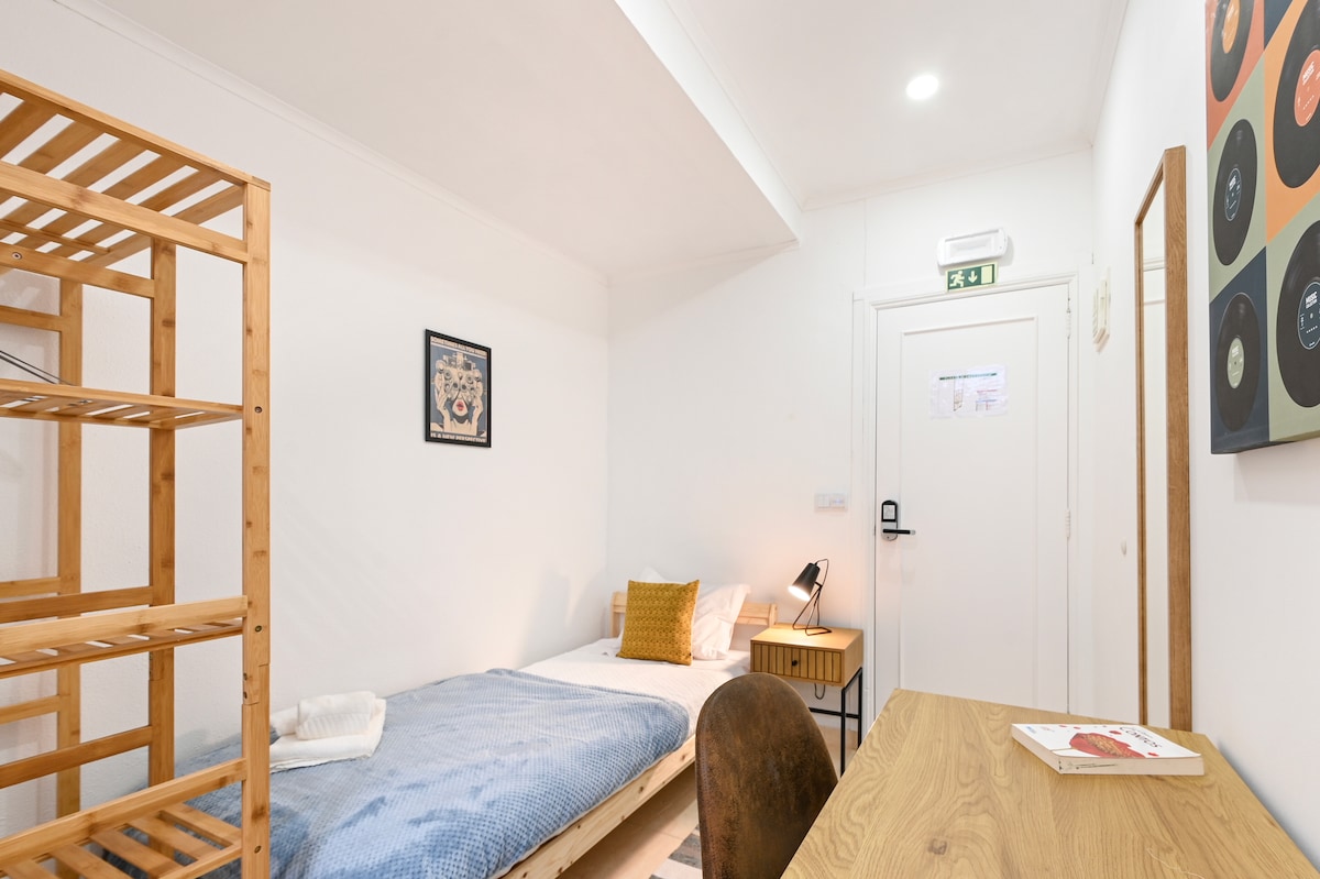 A simple room features a single bed with light blue bedding and a yellow accent pillow. A wooden ladder shelf stands against one wall. A desk with a chair is positioned near the window, which is covered with neutral curtains. A light fixture provides illumination.