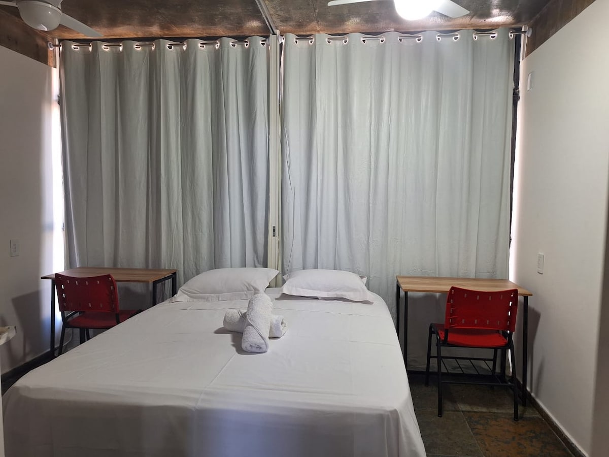 The sleeping area features a spacious bed with two white pillows and a rolled towel placed at the foot. Two simple wooden tables flank the bed, accompanied by red chairs. Light filters gently through sheer curtains, creating a calm and bright environment.