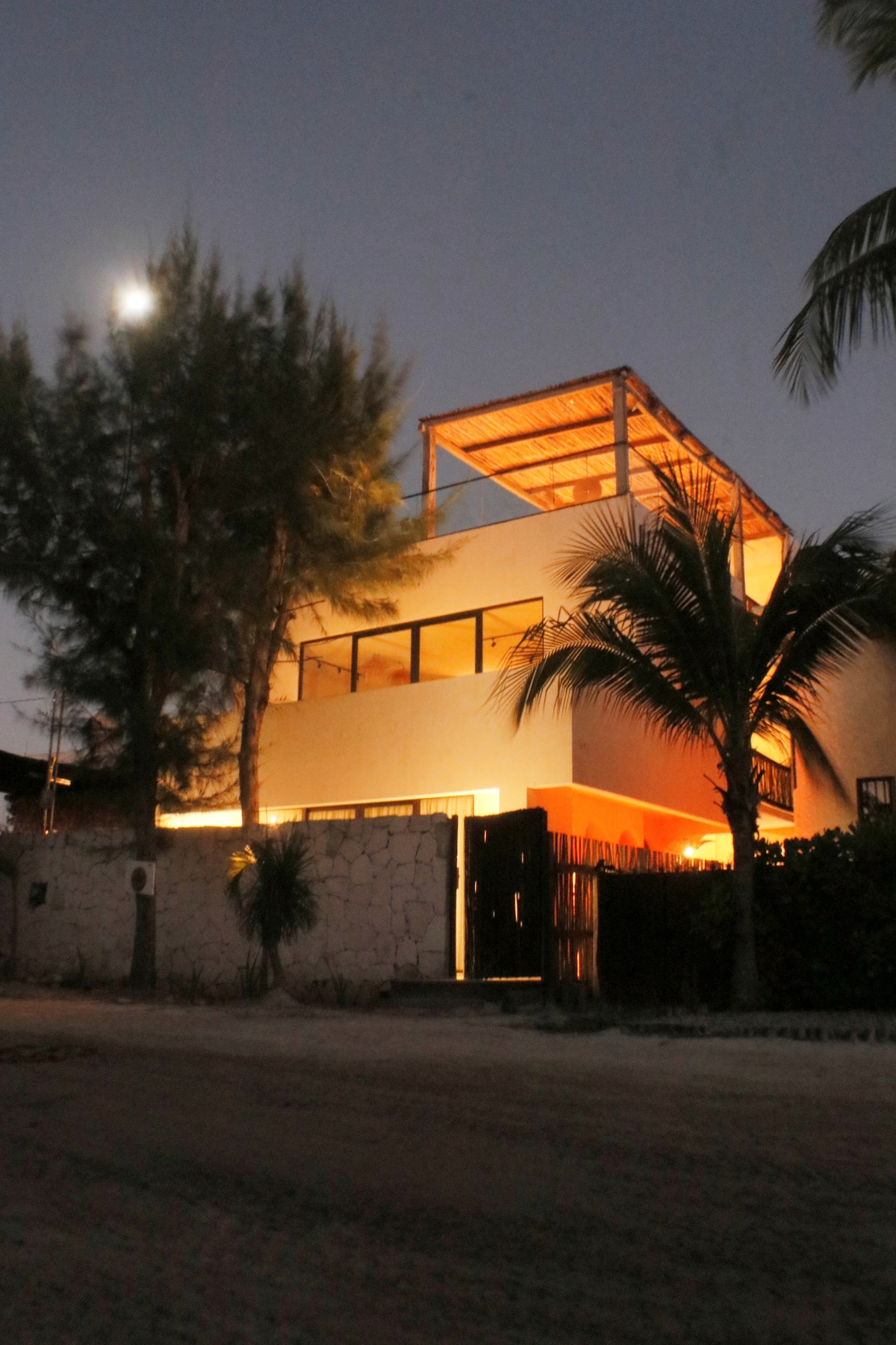 A modern, multi-level building is shown at dusk, featuring a spacious terrace with a straw roof. Soft lighting highlights the structure's clean lines and large windows. Surrounding palm trees enhance the tropical ambiance, while a pathway leads to the entrance, framed by natural stone walls.
