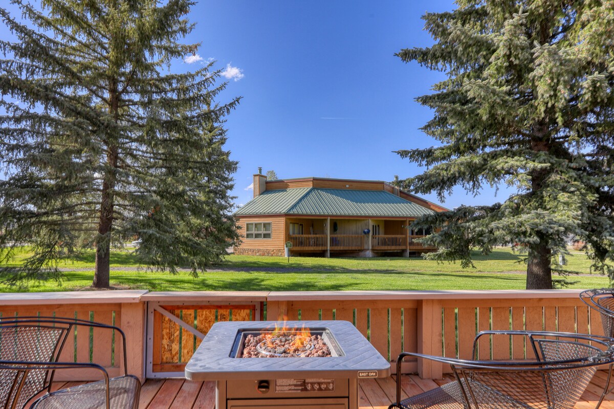 An outdoor deck features a gas fire pit surrounded by comfortable seating. Lush green space is visible in the background, complemented by tall evergreen trees. A spacious building can be seen beyond the trees, enhancing the serene outdoor setting.