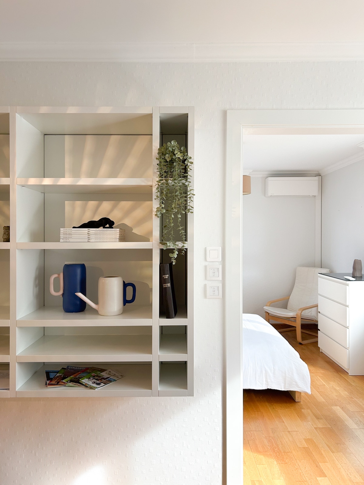 A well-organized shelf is displayed, featuring decorative items such as a blue pitcher, a white vase, and a collection of books. The light from a nearby window casts soft shadows on the shelves. A glimpse of a bright bedroom is visible through the doorway.