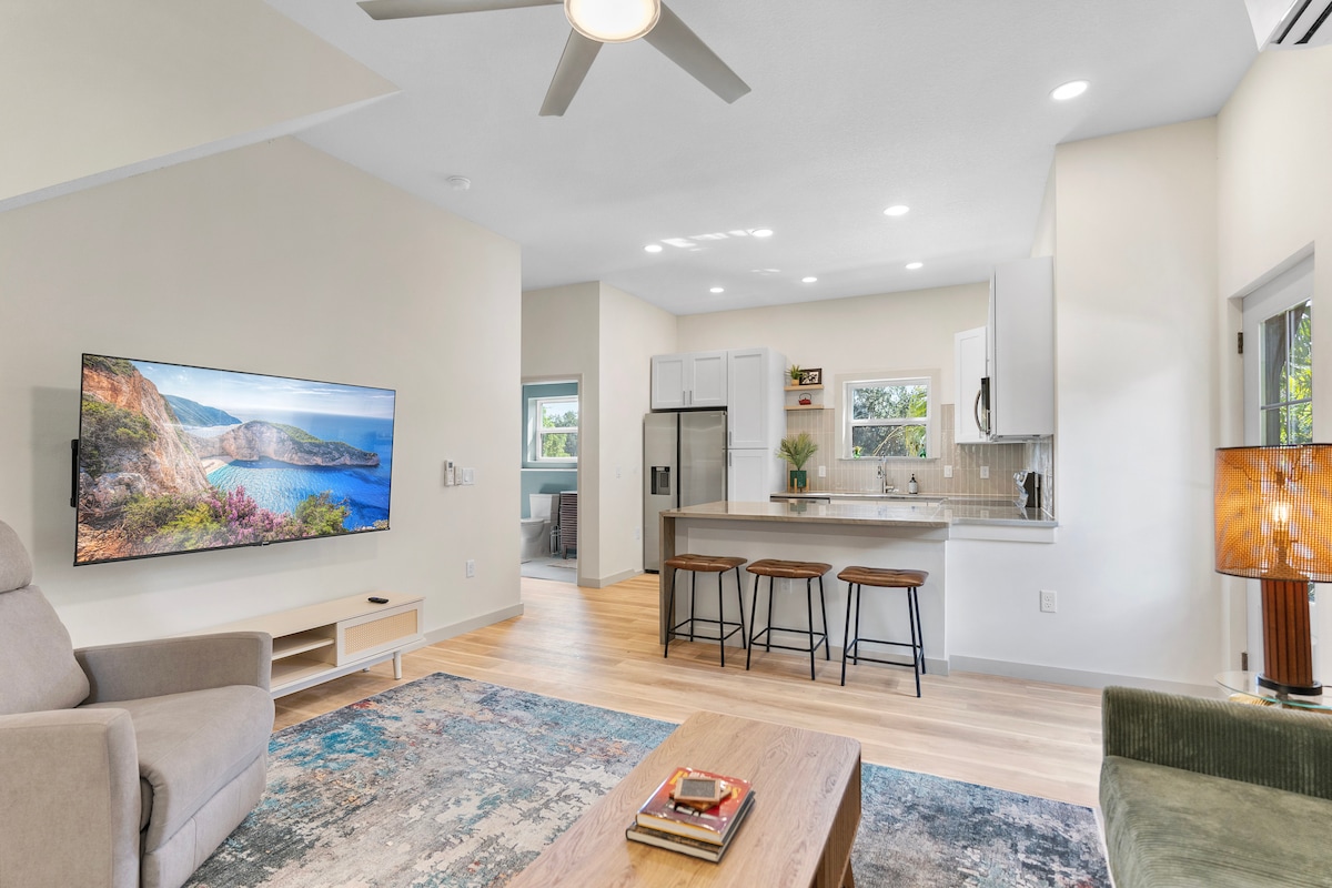 A spacious living area features a large flat-screen TV mounted on a pale wall. A comfortable sofa and an armchair provide seating, while a wooden coffee table sits in the center. Natural light fills the space, highlighting the open kitchen with modern appliances and a breakfast bar.