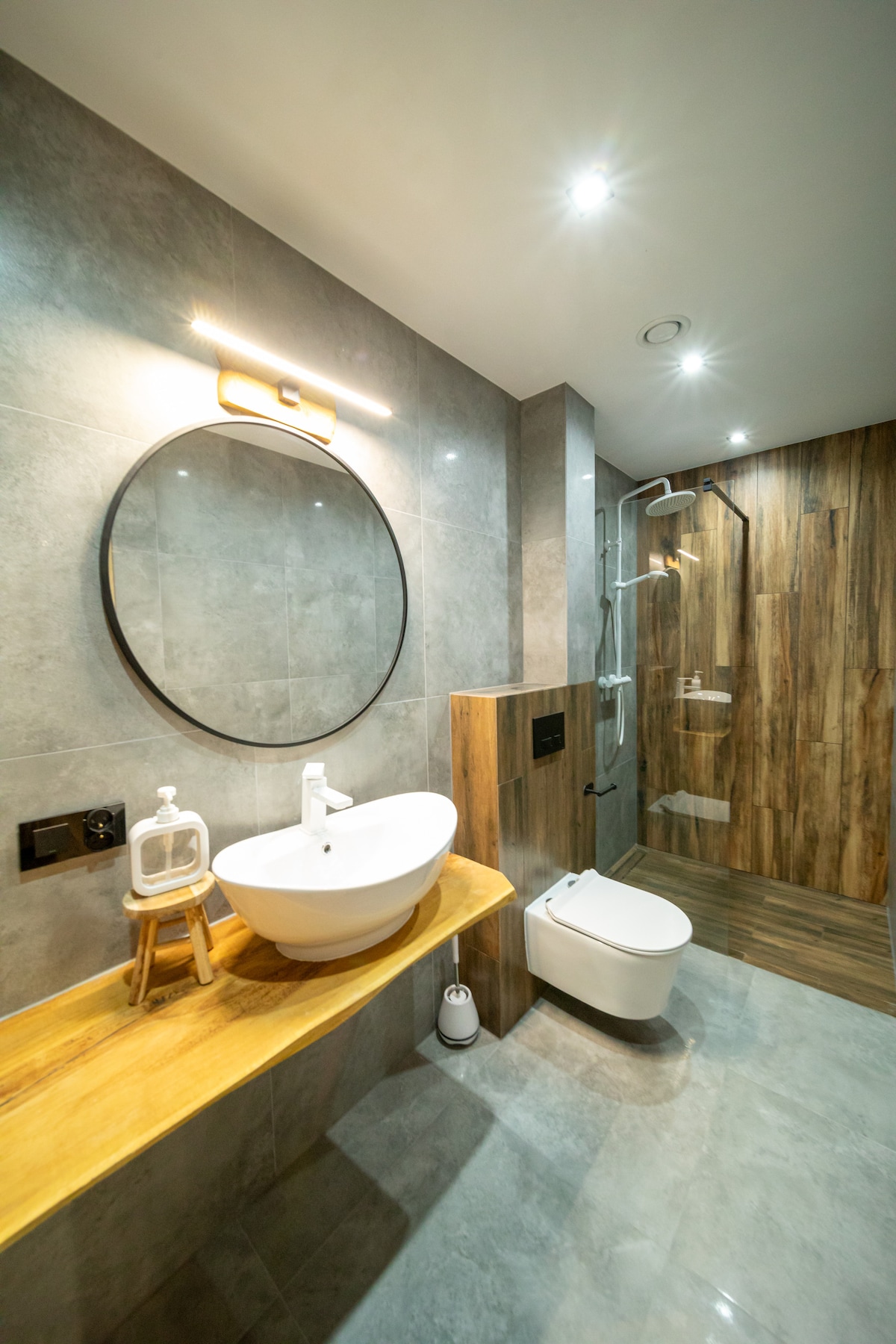 A modern bathroom features elegant gray tiles and wooden accents. A round mirror hangs above a stylish sink set on a wooden countertop. A glass shower enclosure and a wall-mounted toilet are visible, complemented by softly illuminated spotlights in the ceiling.